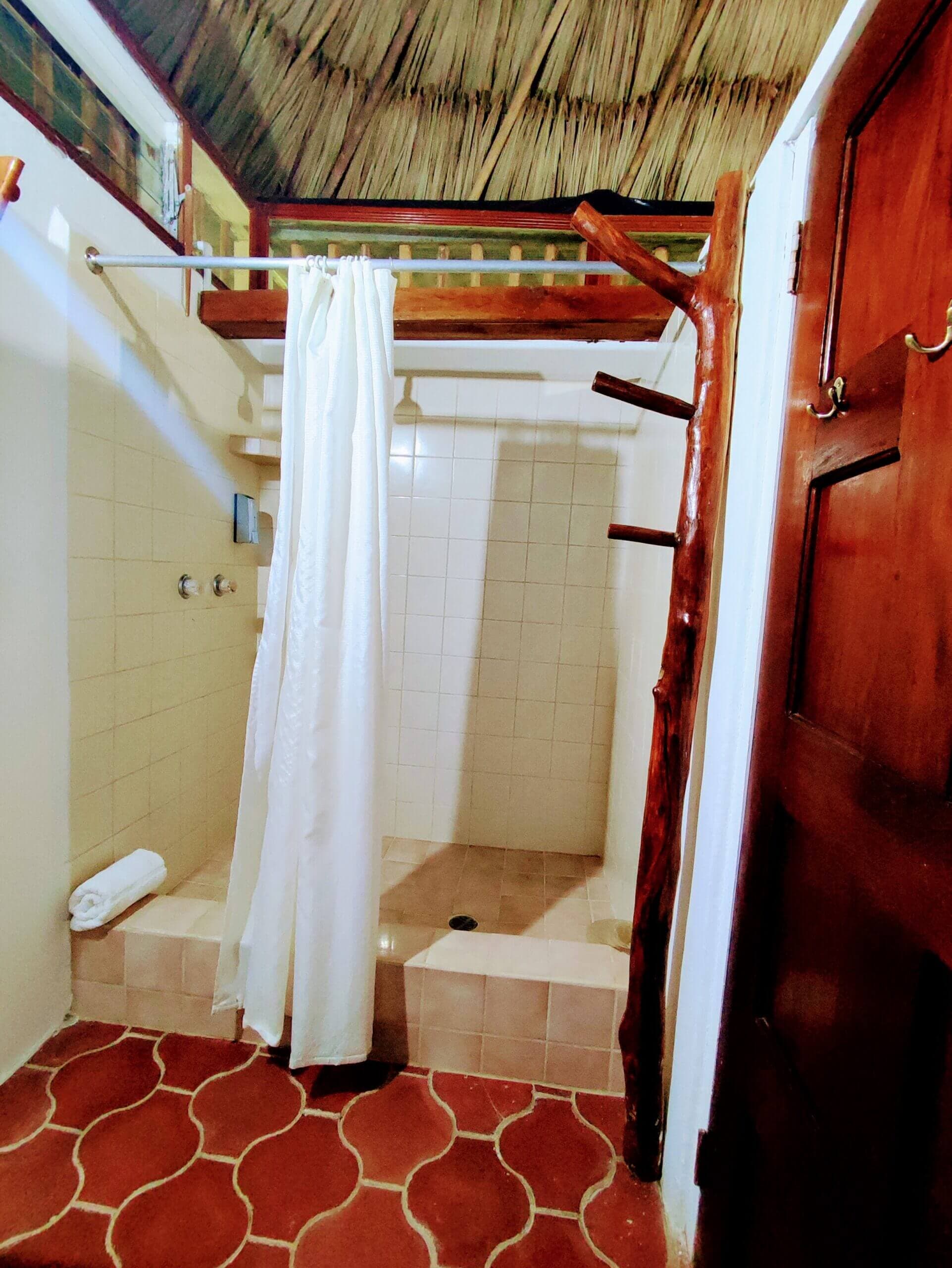 A shower area with a white curtain, tan-tiled walls, and red patterned tile flooring, located beneath a high thatched roof with wooden structural beams.