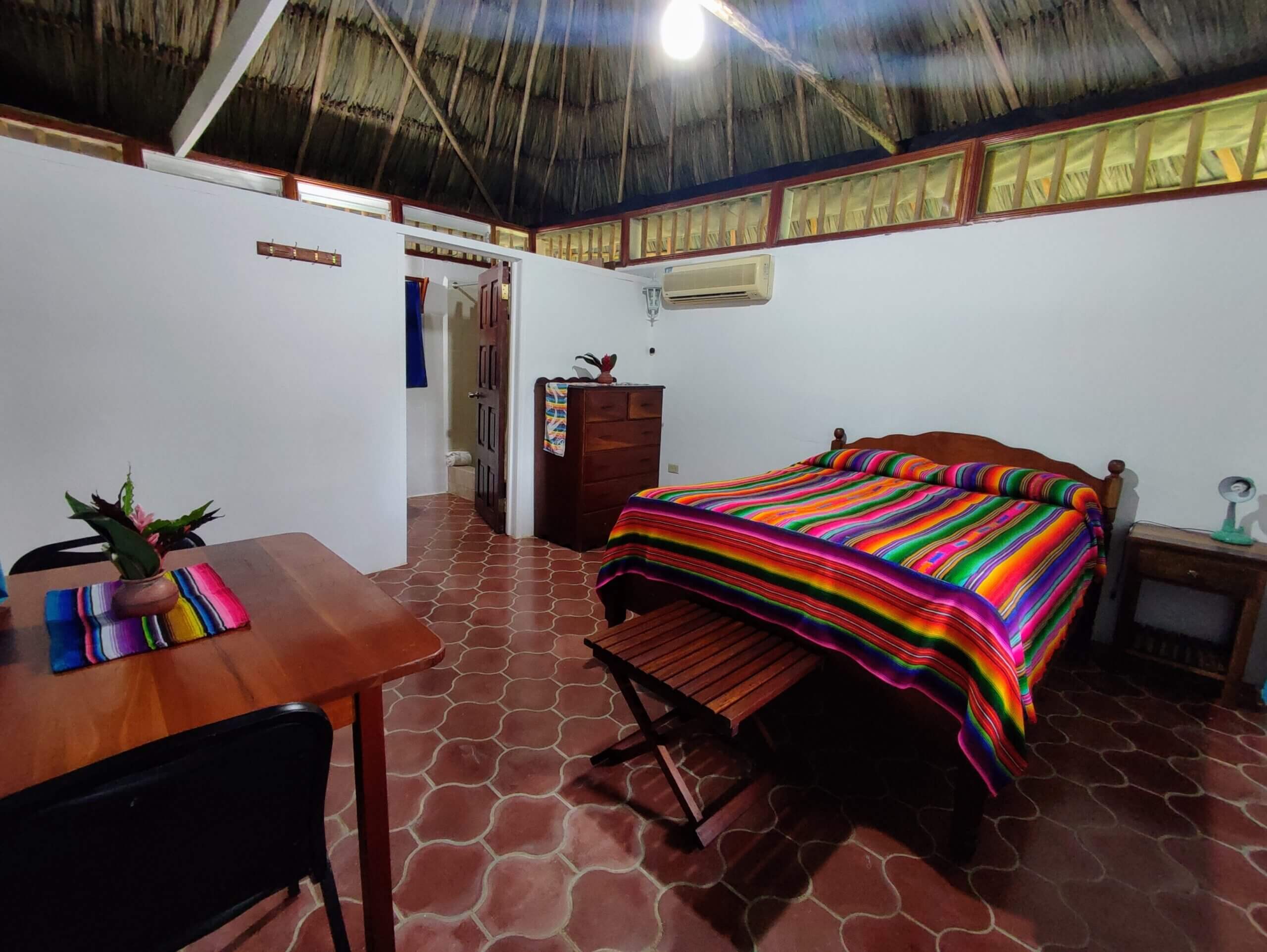 A bedroom with white walls and a thatched roof, featuring a bed covered in a vibrant multi-colored striped blanket, a small wooden table, and red tile flooring.