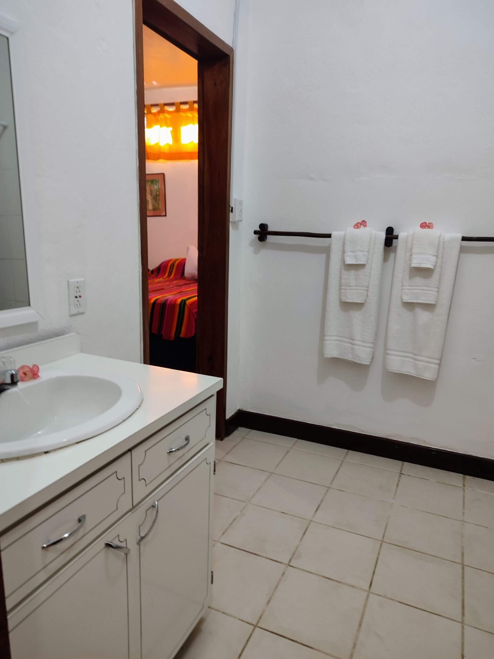 A simple bathroom with white walls and light-colored tile flooring, featuring a white vanity with a sink and white towels hanging on a dark wood rack, with a view through an open doorway into a bedroom.