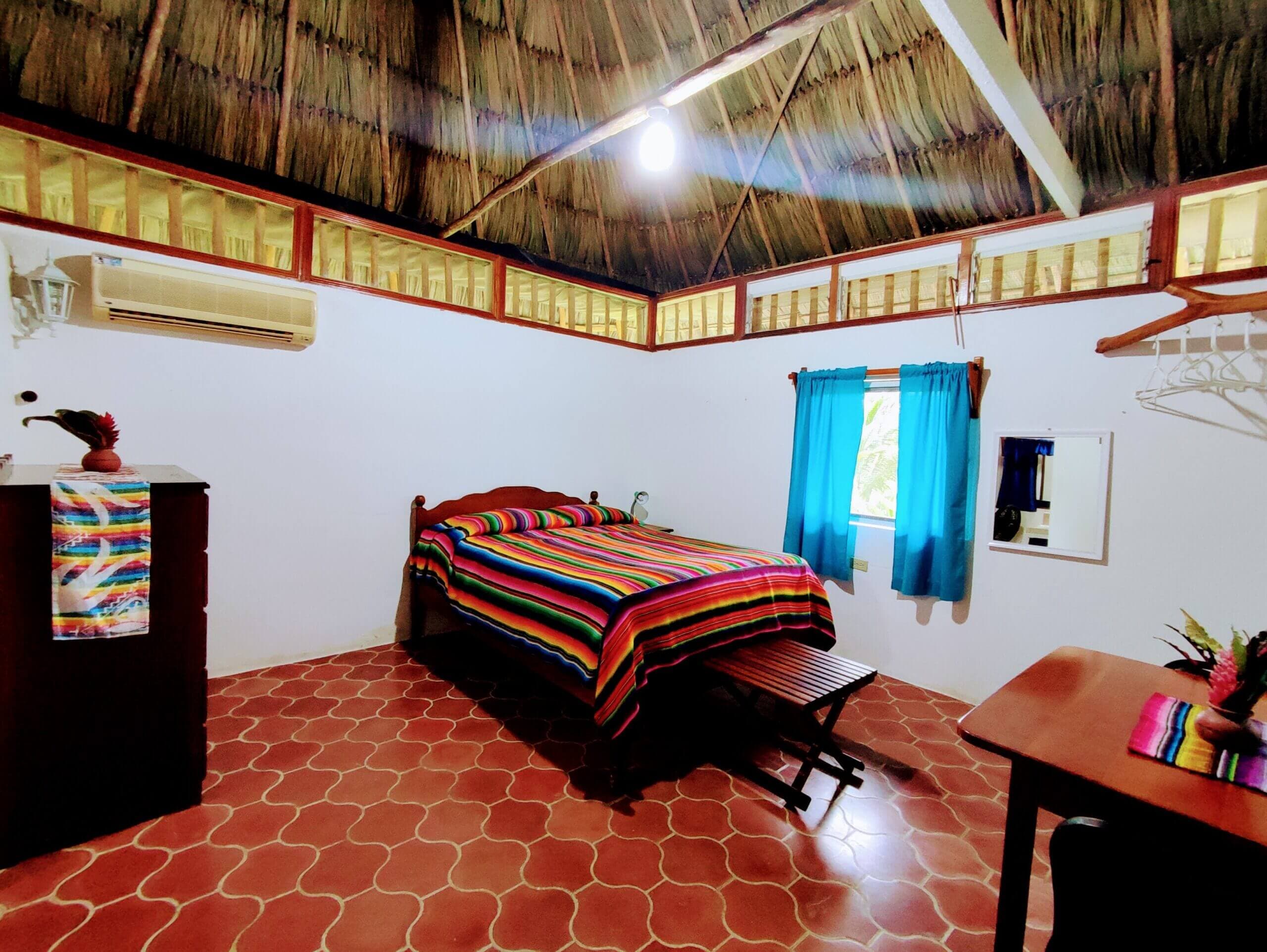 A bedroom with white walls, a thatched roof, and red tile flooring, featuring a bed with a vibrant multi-colored striped blanket and a small window with blue curtains.