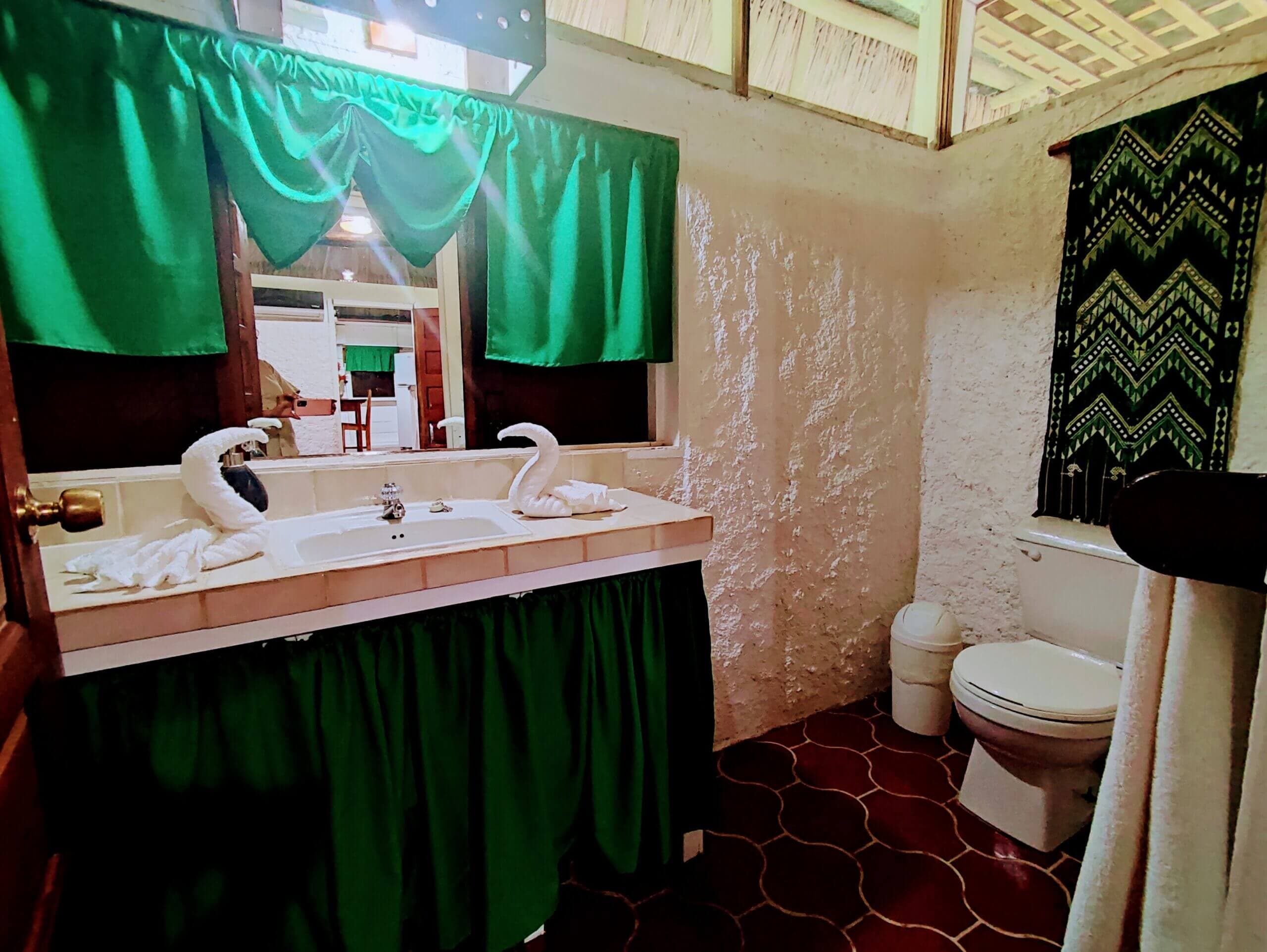 A simple bathroom with red patterned tile flooring, featuring a white pedestal sink with green fabric covering the base, a toilet, and a large mirror framed by bright green curtains.