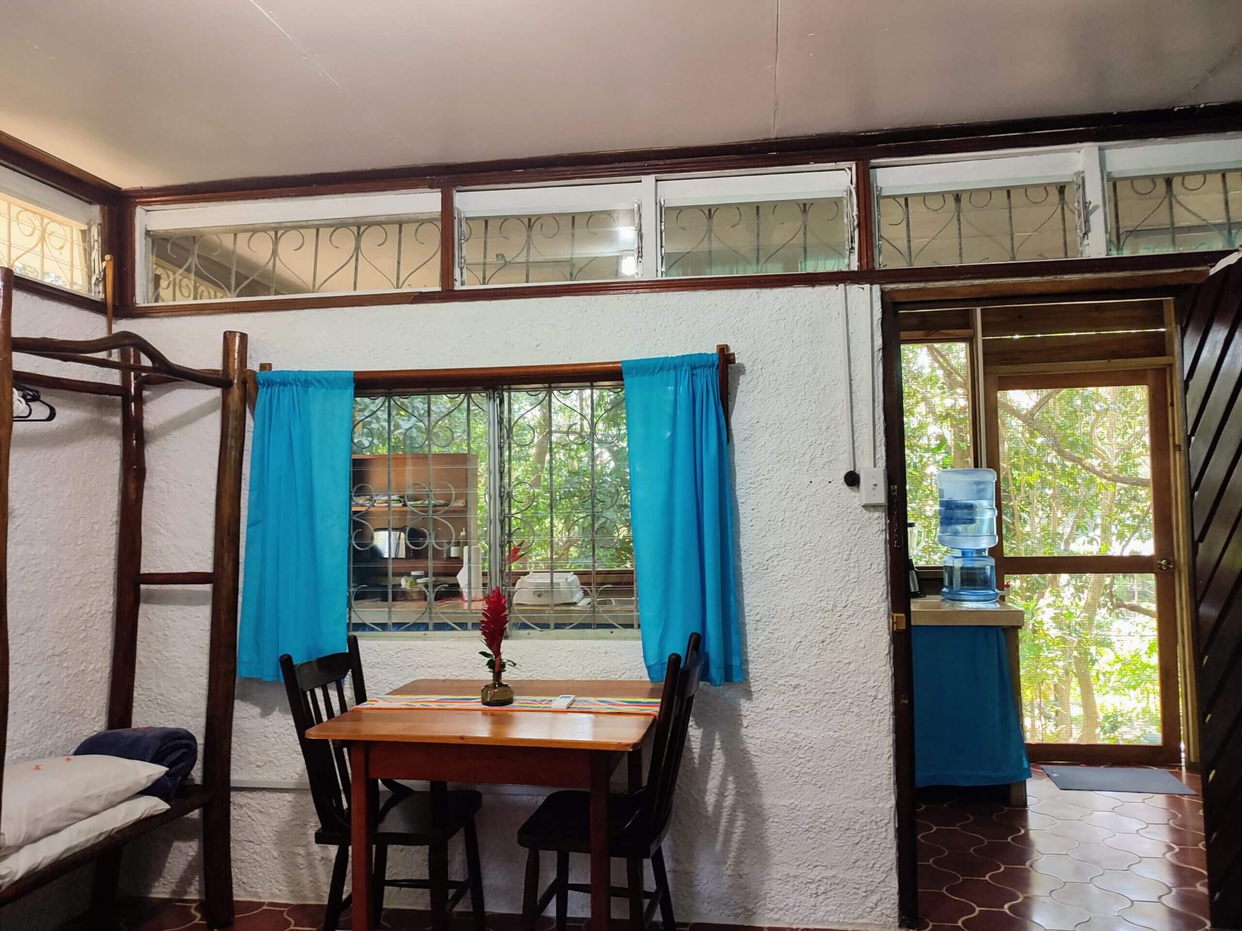A dining area with white walls and red patterned tile flooring, featuring a wooden table with black chairs situated near a window with bright teal curtains, with a view through a doorway into a kitchenette with a blue fabric-skirted counter.