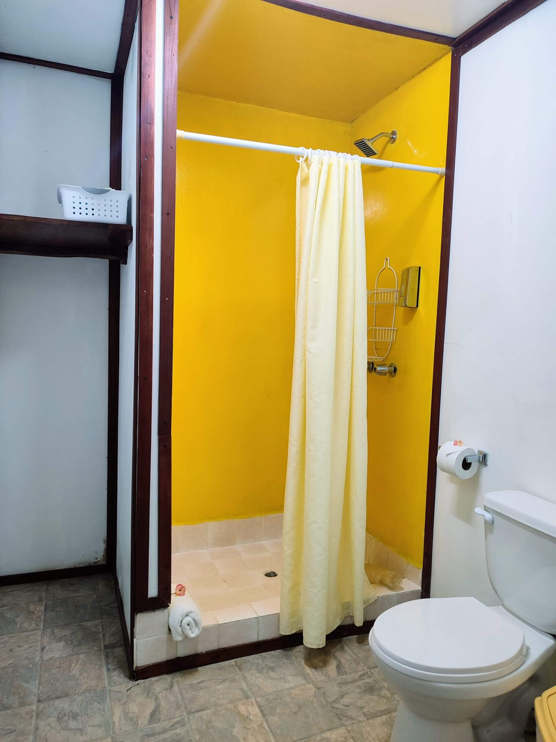 A bathroom featuring a shower stall with bright yellow walls and a pale yellow curtain, a white toilet, and light-colored tile flooring.