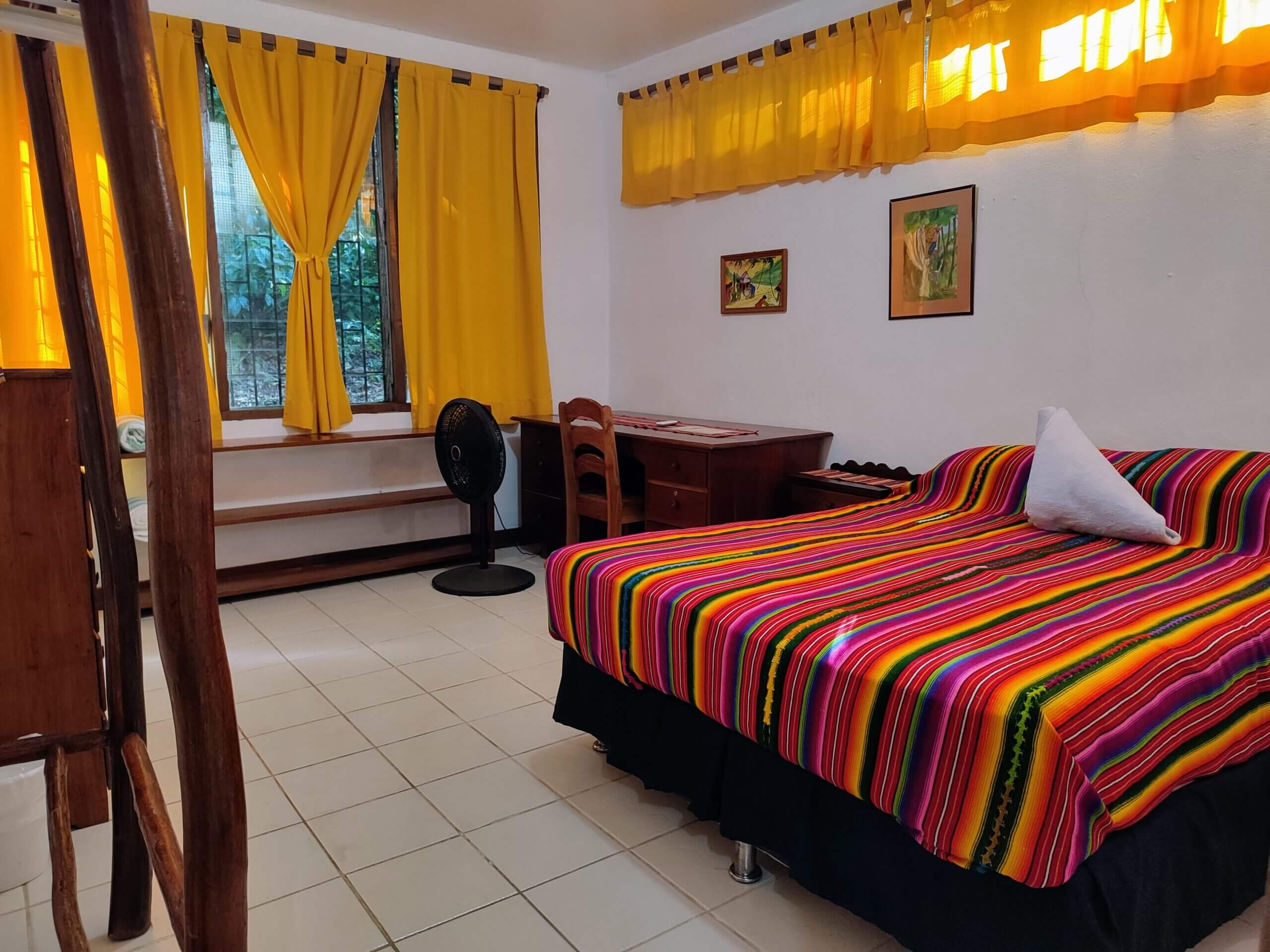 A bedroom with white walls and light-colored tile flooring, featuring a large bed with a vibrant multi-colored striped blanket, a wooden desk with a chair, and windows with bright yellow curtains.