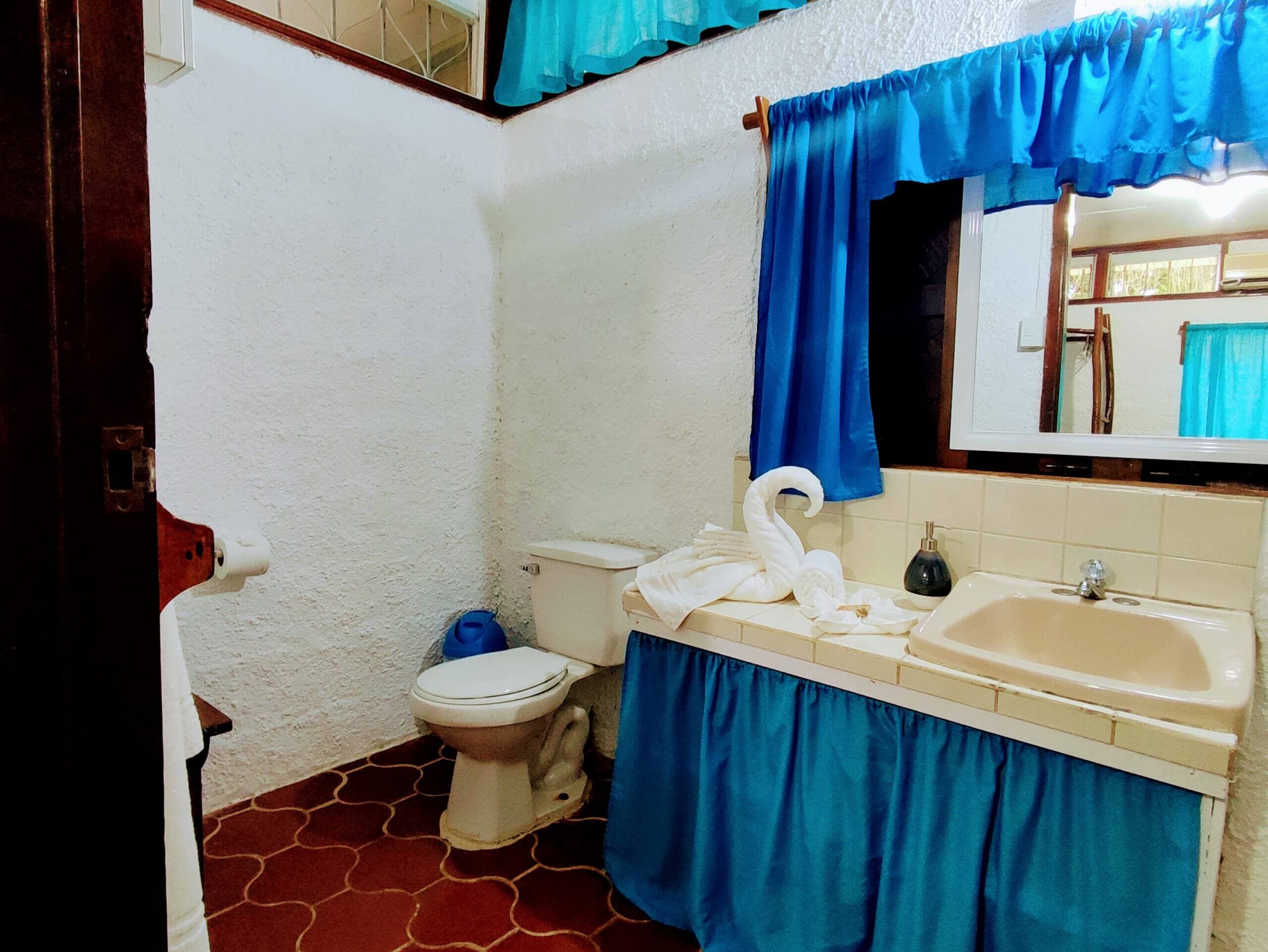 A bathroom featuring a white vanity with a bright blue fabric skirt and matching blue curtains over a mirror, a white toilet, and red patterned tile flooring.