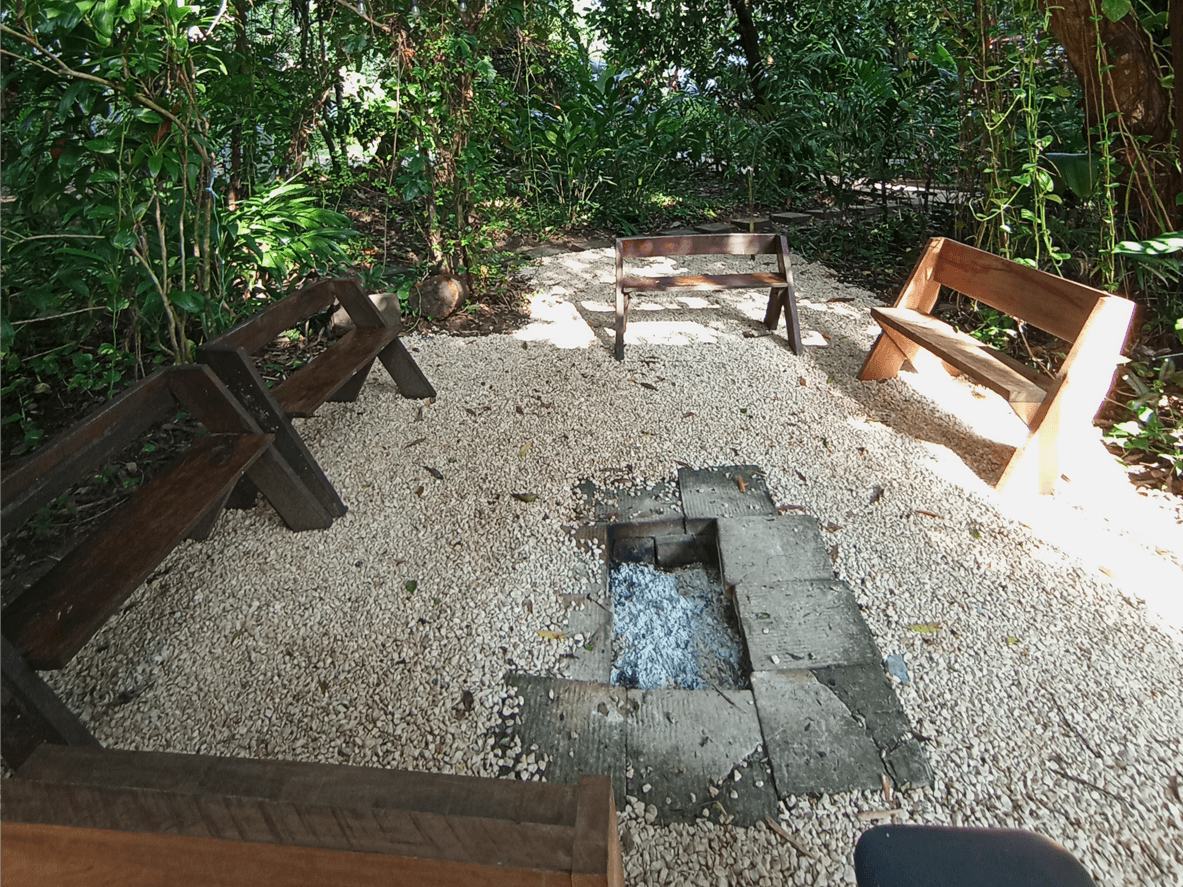 A secluded outdoor seating area surrounded by greenery, featuring wooden benches arranged around a stone fire pit.