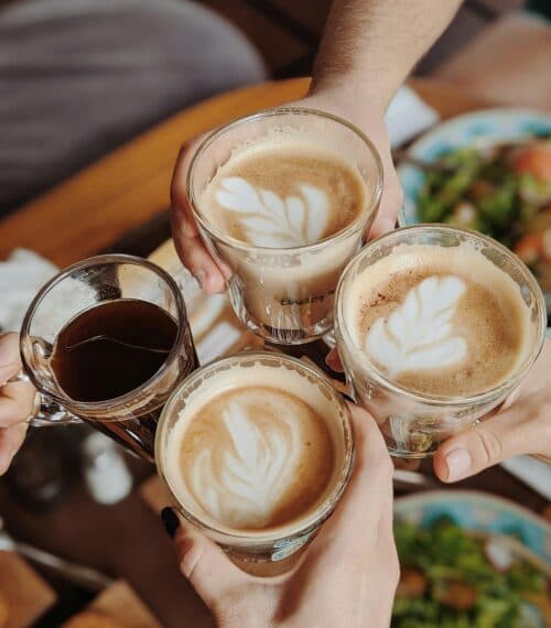 Four hands holding coffee cups with latte art, toasting over a wooden table.