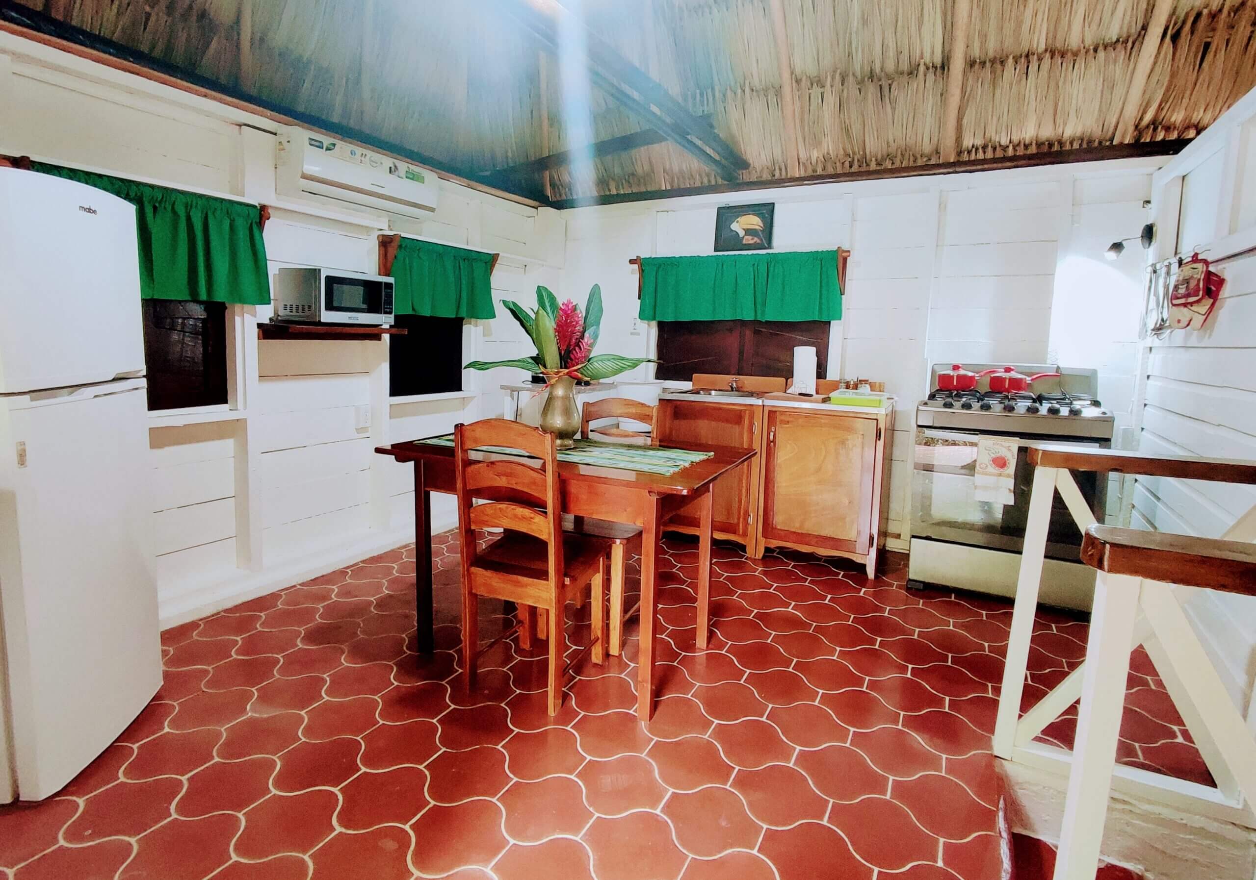 An open-concept kitchen and dining area with red patterned tile flooring, featuring a wooden dining table, a white refrigerator, a black stove, and windows with bright green curtains under a vaulted ceiling.