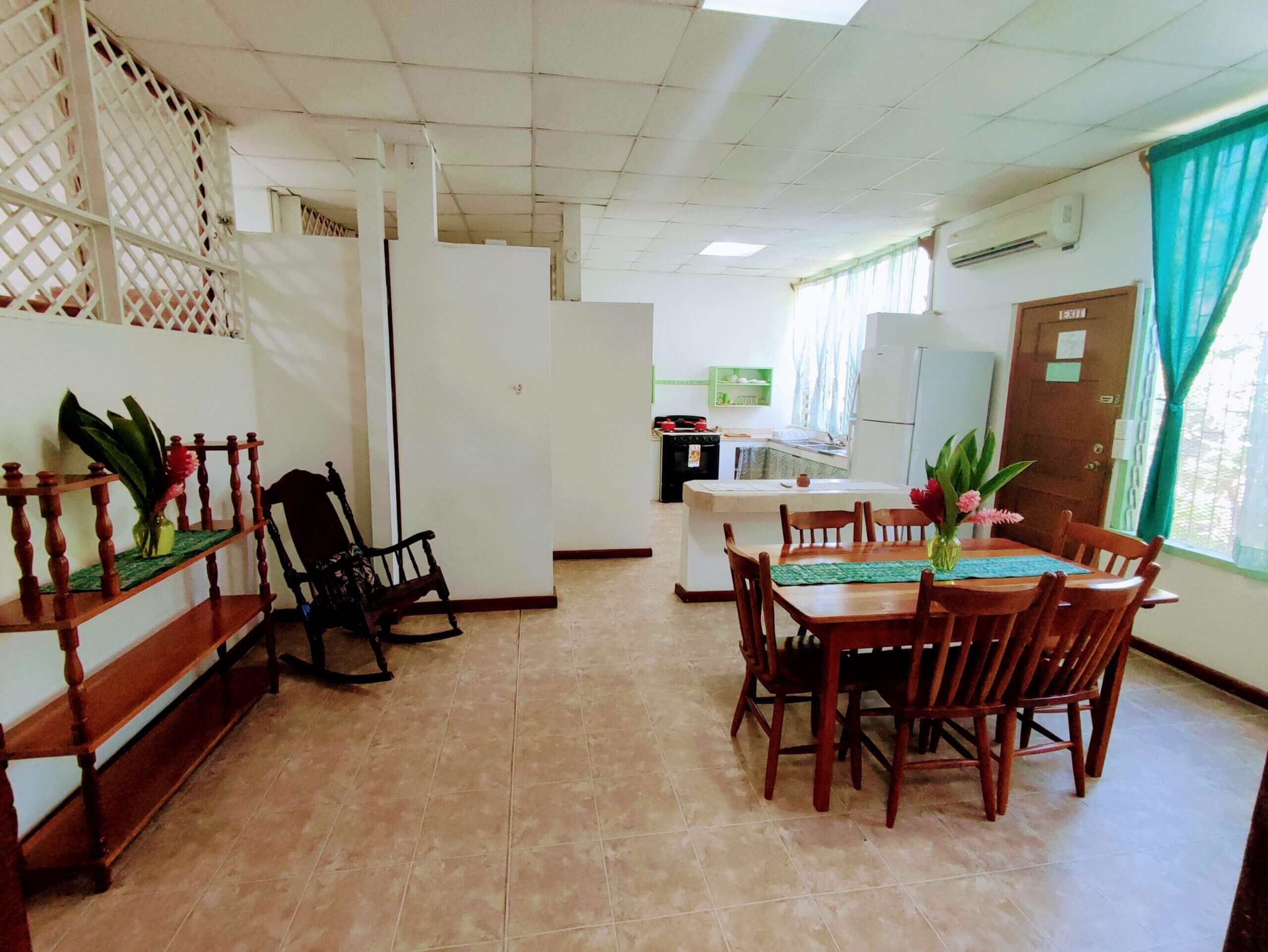 A spacious dining and living area with light-colored tile flooring, featuring a wooden dining table with chairs and a black rocking chair, with a view into a white kitchen area in the background.