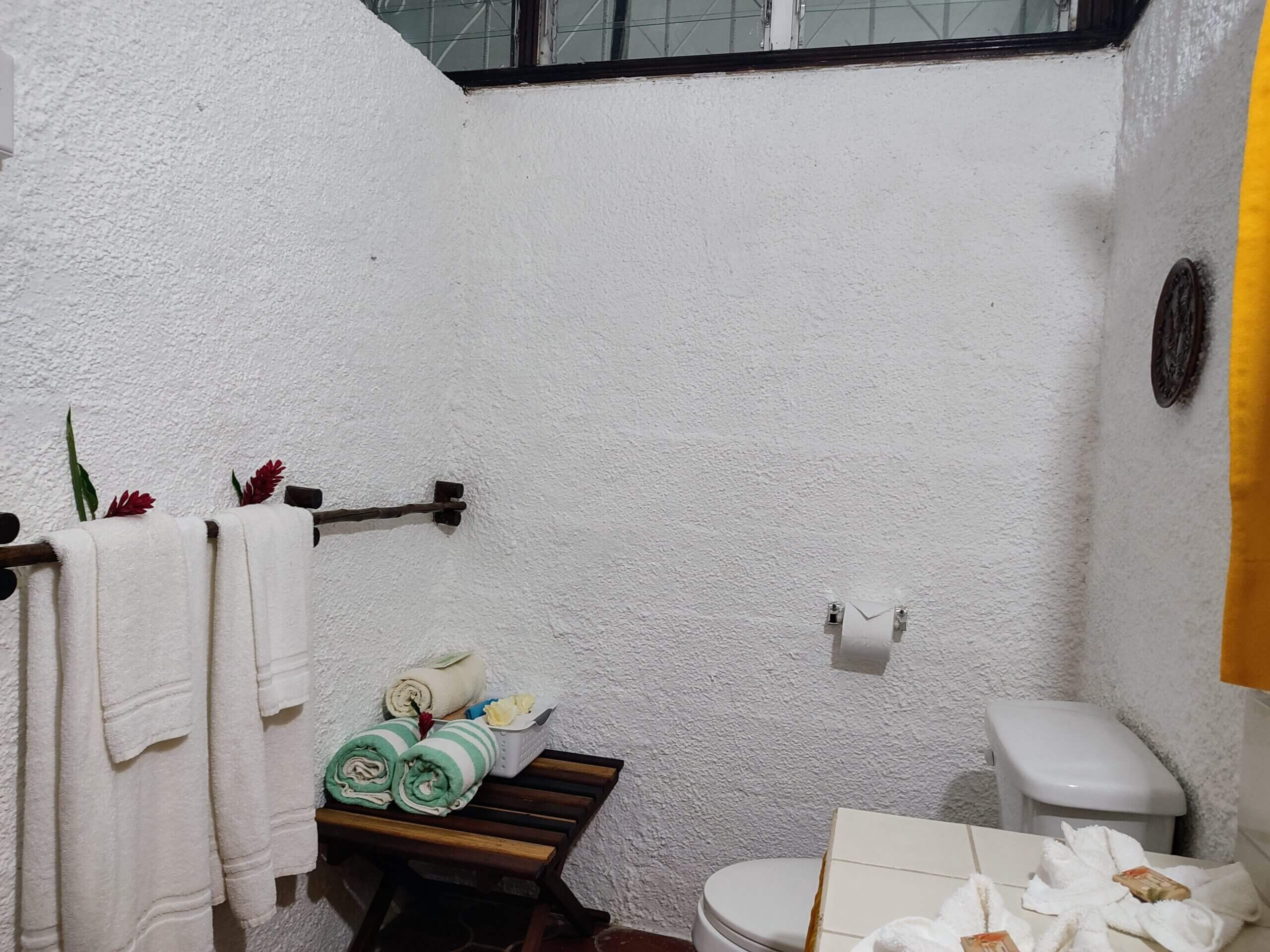 A bathroom area with white brick-textured walls, featuring a wooden bench stacked with rolled light-green towels, white hand towels hanging on a dark metal rack, and a white toilet.