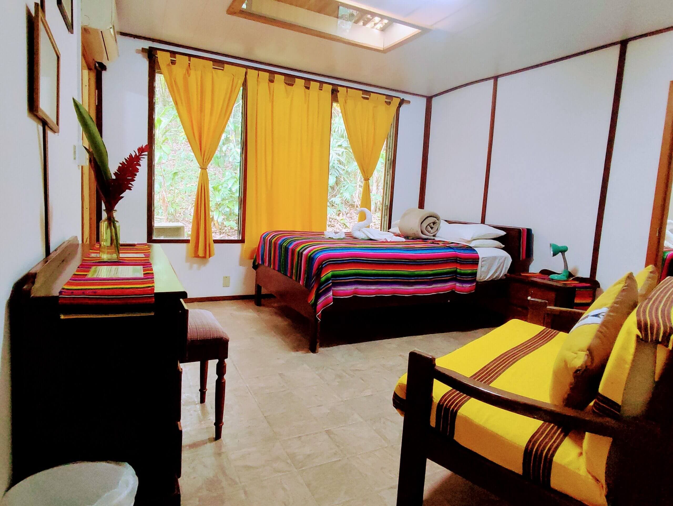 A bedroom featuring white paneled walls and light-colored tile flooring, with a bed covered in a vibrant multi-colored striped blanket and a wooden sofa with yellow cushions positioned near a window with yellow curtains.