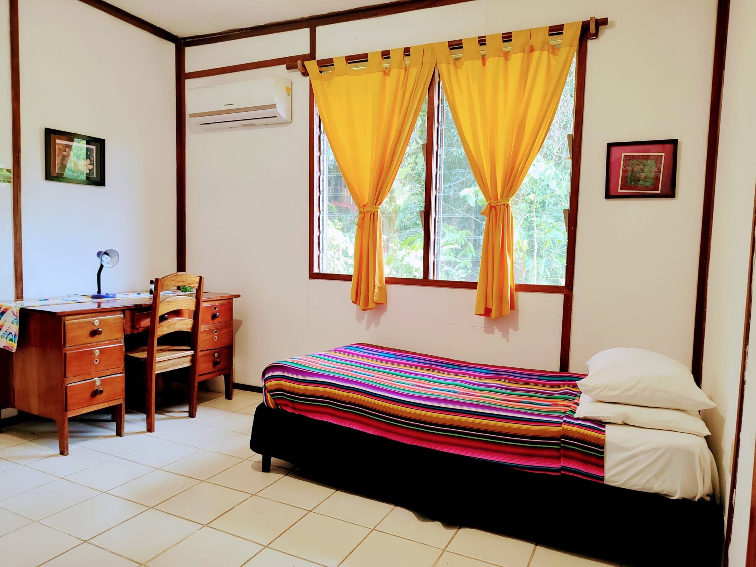 A bedroom with white walls and light-colored tile flooring, featuring a single bed with a vibrant multi-colored striped blanket and a wooden desk with a chair beneath a window with bright yellow curtains.