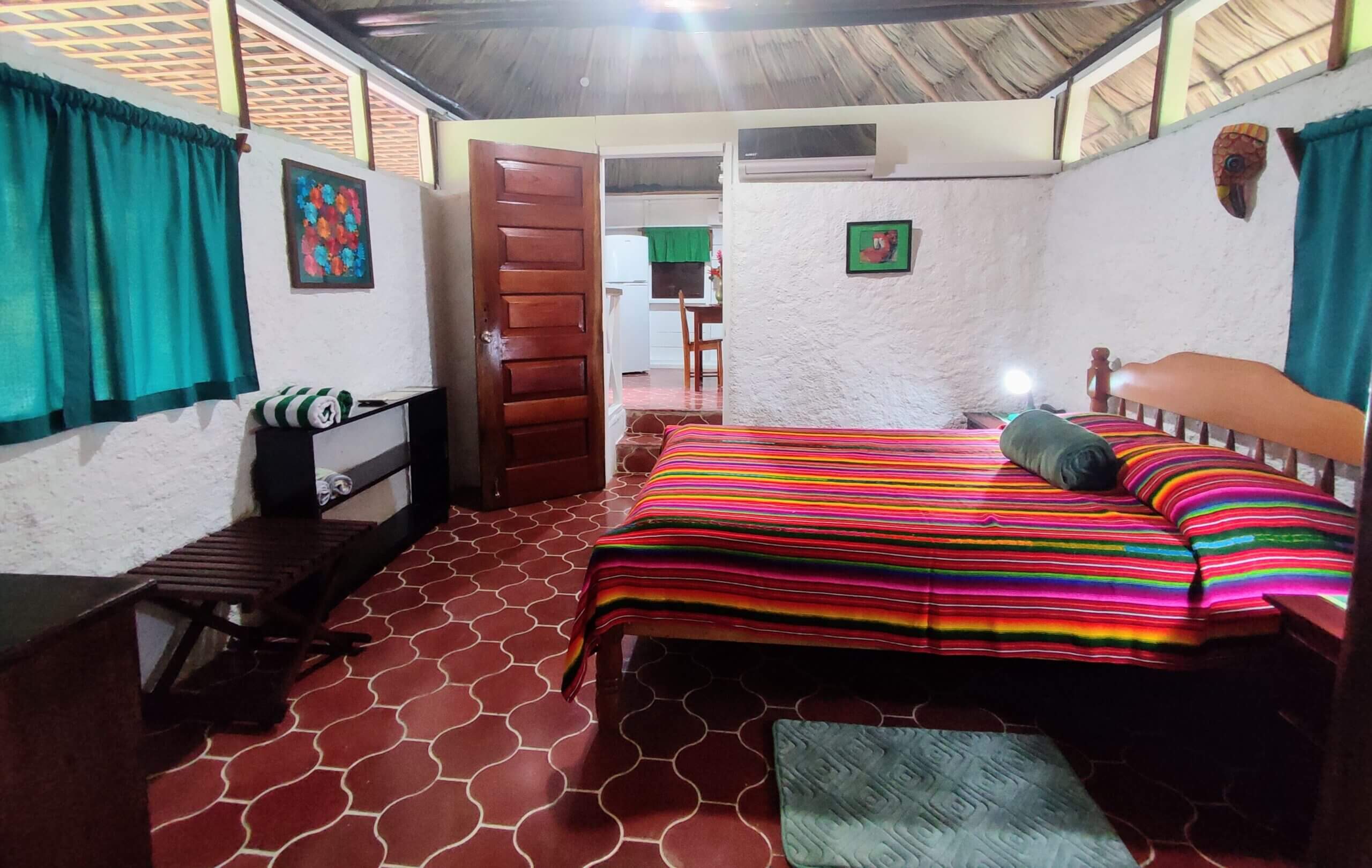 A bedroom with red patterned tile flooring and a vaulted wooden ceiling, featuring a large bed with a vibrant multi-colored striped blanket, dark wood furniture, and windows with deep green curtains.