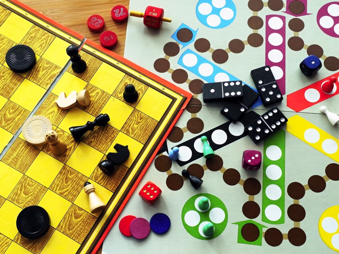 A variety of board games, including chess, ludo, and dominoes, are scattered on a wooden table.
