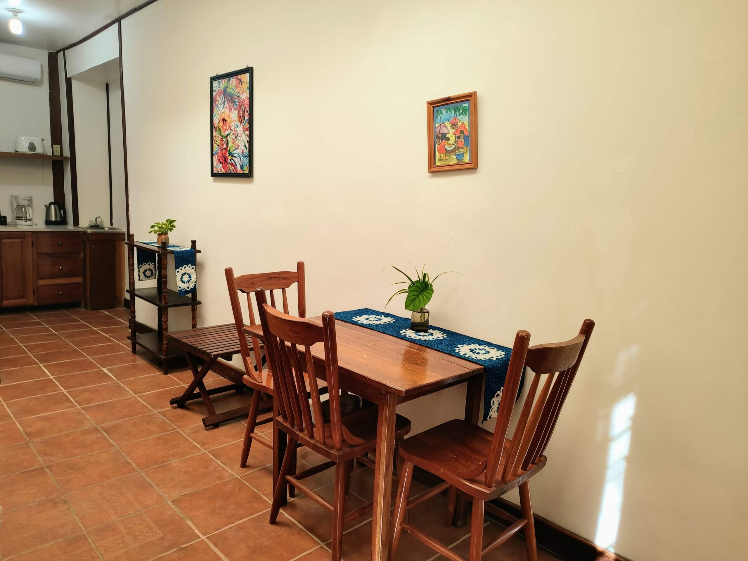 A simple dining area with terra cotta tile flooring, featuring a small wooden table with three matching chairs set against a plain cream-colored wall decorated with two framed pictures.