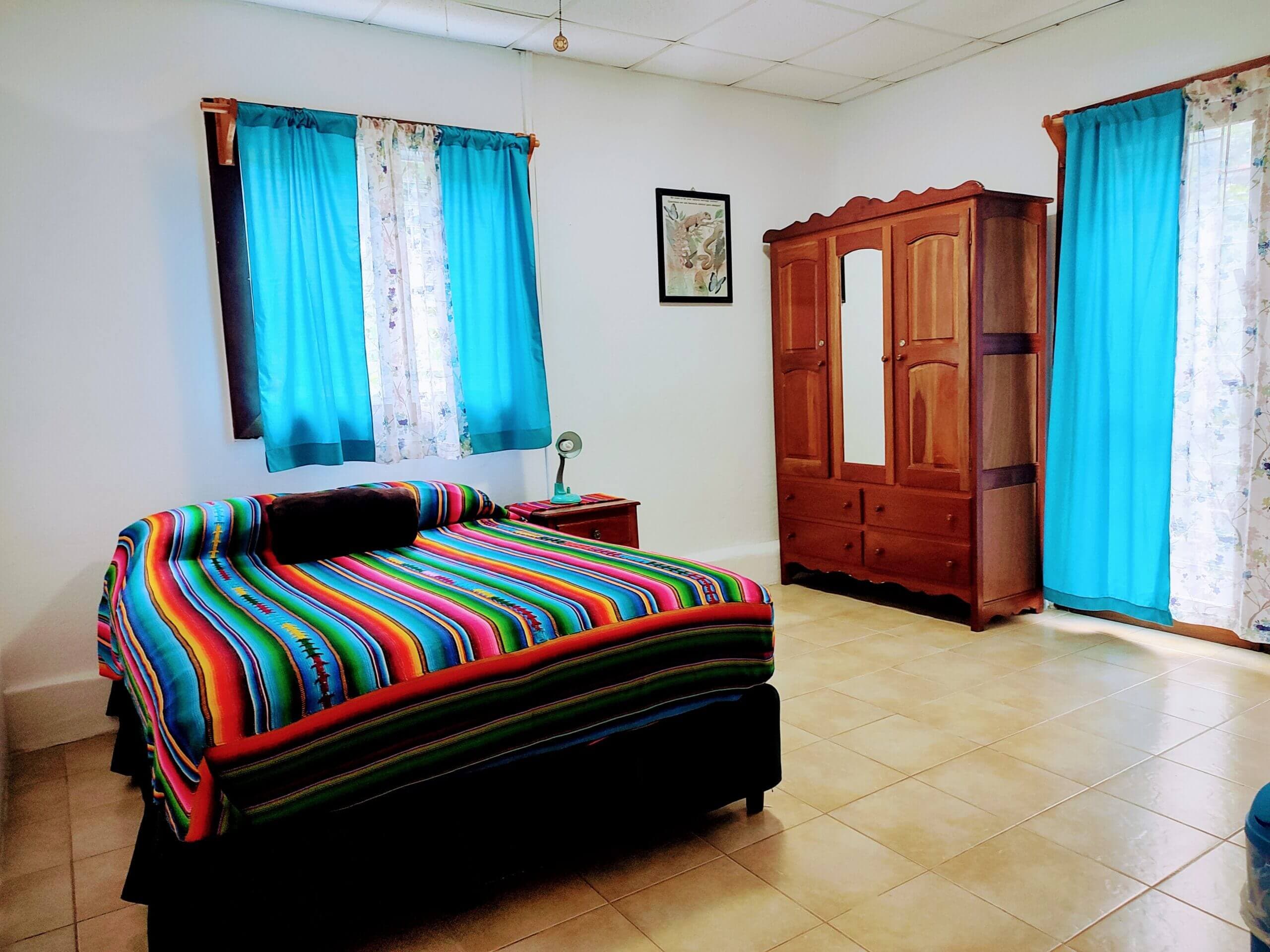 A spacious bedroom with white walls and light-colored tile flooring, featuring a large bed with a vibrant multi-colored striped blanket, a tall wooden wardrobe with a mirror, and windows with bright teal curtains.