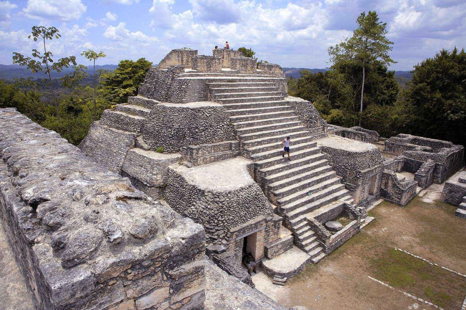 A stone pyramid structure with stepped terraces surrounded by lush greenery under a blue sky.