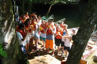 A group of children in life vests gathers by the water, holding snacks and smiling.