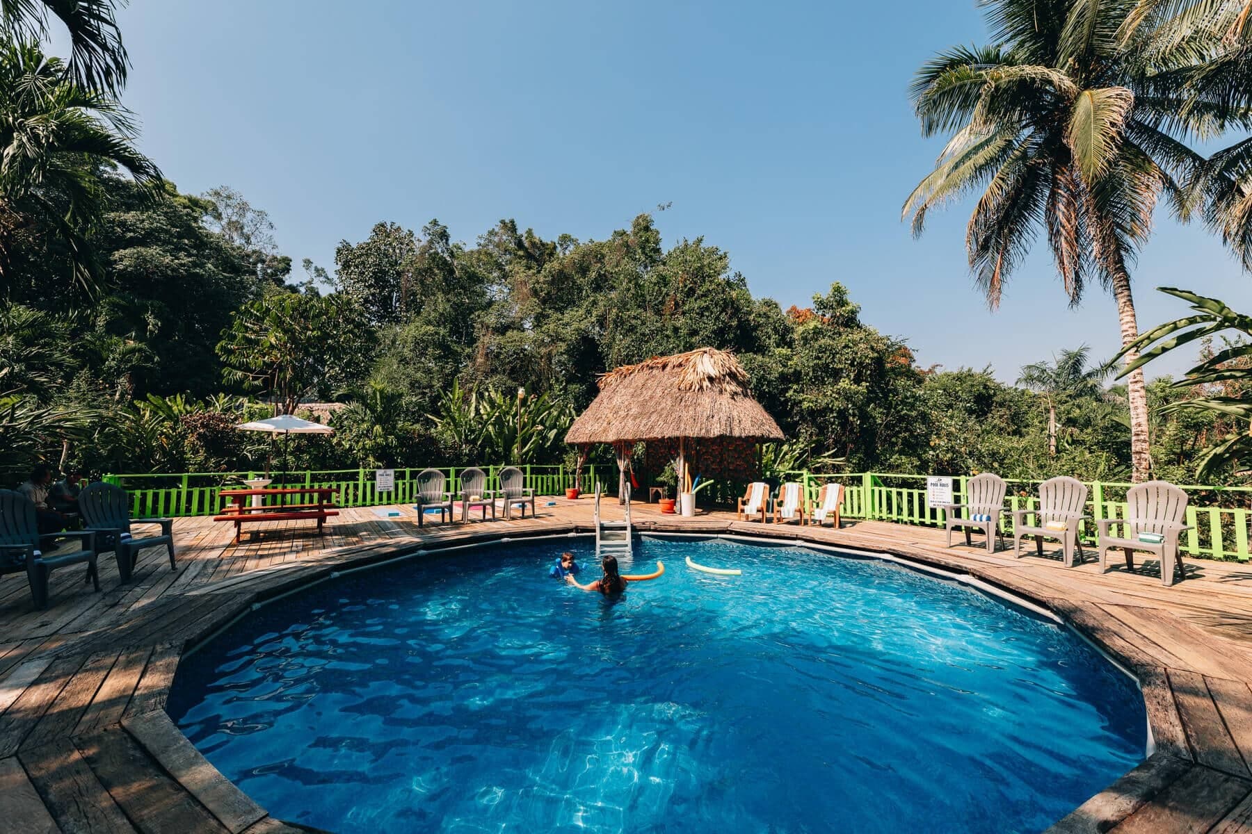 A blue swimming pool surrounded by tropical greenery and seating areas, featuring a thatched gazebo.