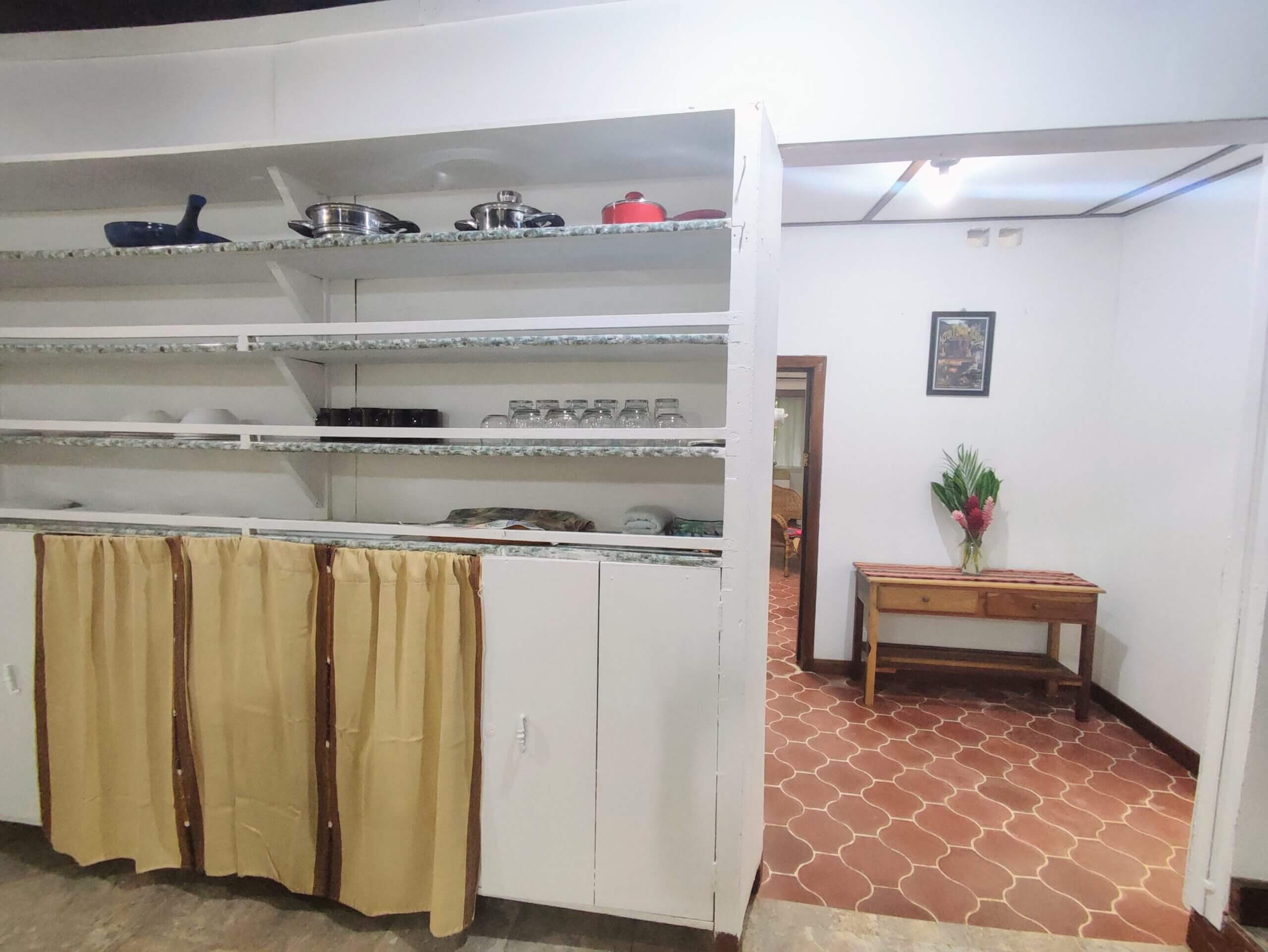 A white storage unit with built-in shelving and tan fabric curtains covering the lower cabinets, positioned next to a doorway that reveals a room with red patterned tile flooring and a small wooden table.