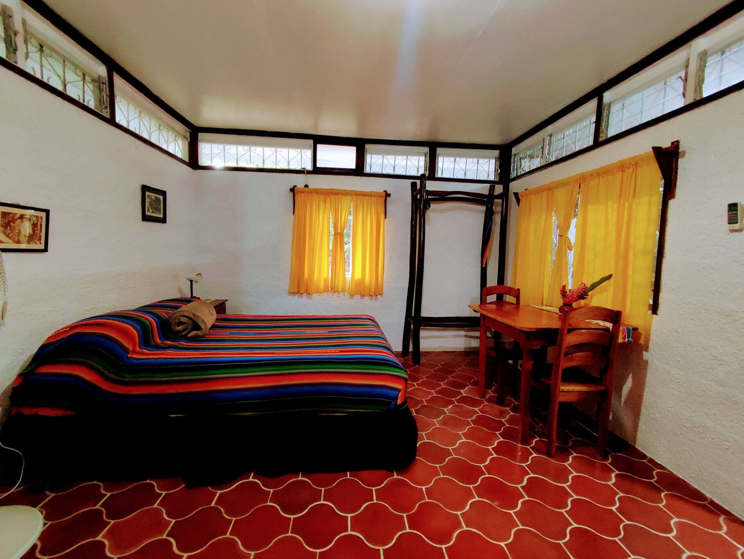 A bedroom with white walls and red patterned tile flooring, featuring a bed with a vibrant multi-colored striped blanket, a small wooden dining table with chairs, and windows with bright yellow curtains.